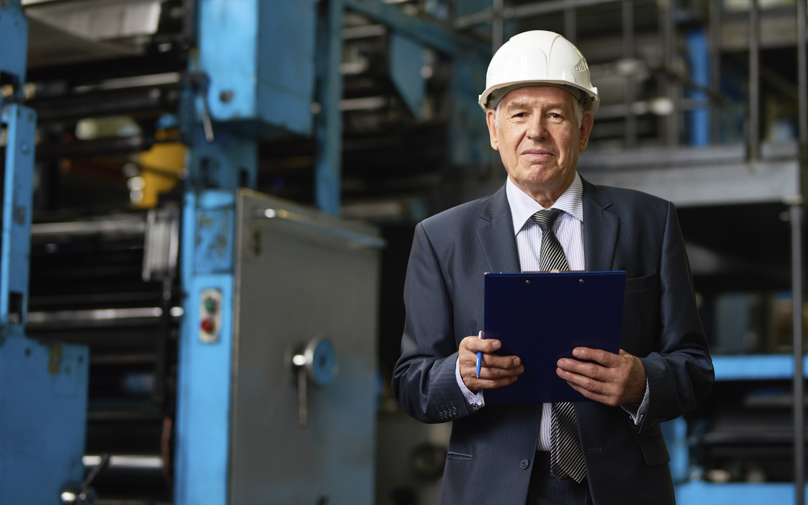 Bauleiter steht mit Anzug, Helm und Unterlagen in einer Industrie