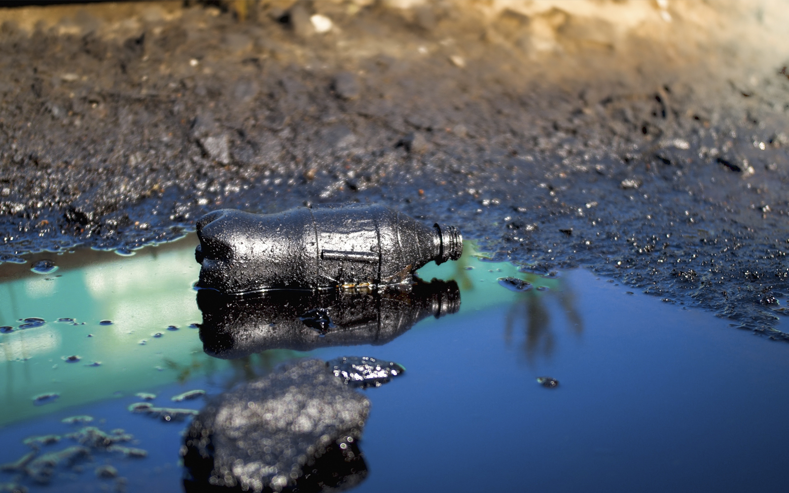 Schwarzes, verschmutzes Wasser in einer Pfütze, in welchem eine verdreckte Plastikflasche liegt