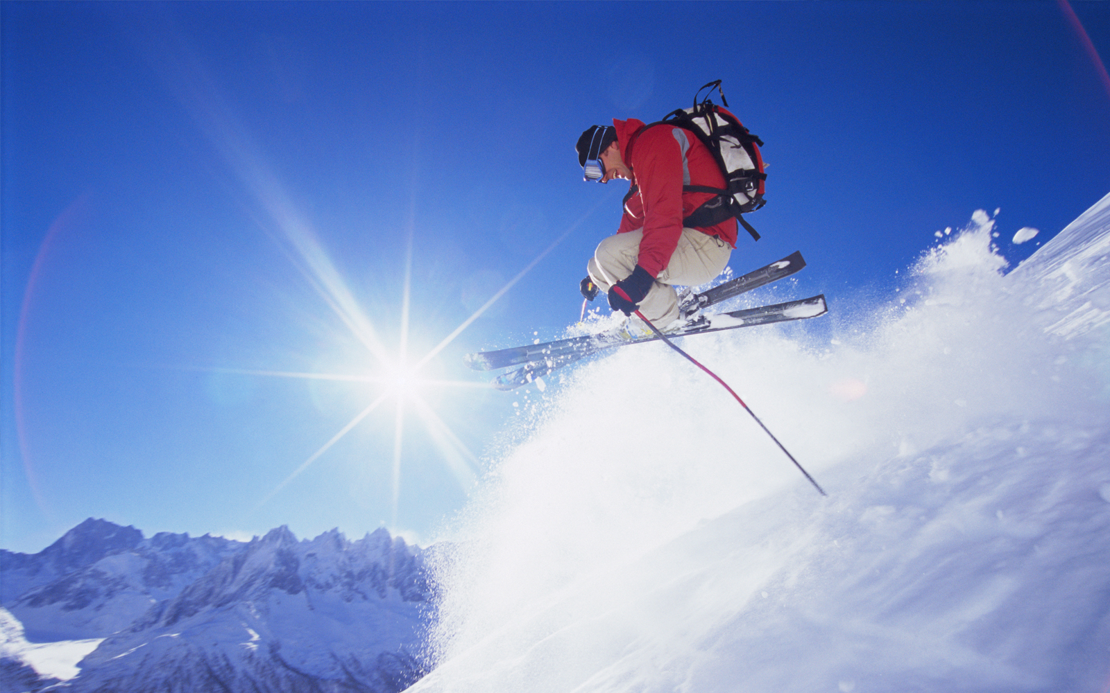 Mann welcher bei strahlendem Wetter Ski fährt. Im Hintergund sind die Berge zu sehen. Der Mann schwebt gerade mit den Ski in der Luft und lacht.
