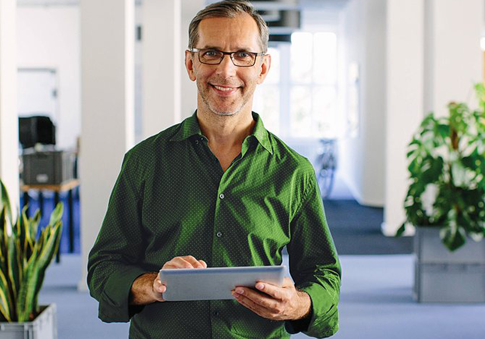 Mann mit einem grünen Hemd steht in einem Büro, lacht in die Kamera und notiert etwas auf Schreibblock